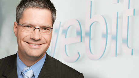 Ein lächelnder Mann mit Anzug, Schlips und Brille, im Hintergrund der webit! Schriftzug