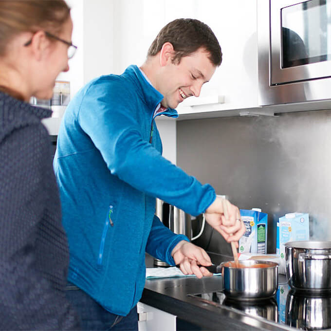 Ein Mitarbeiter rührt einen Kochtopf mit Tomatensoße, eine Mitarbeiterin sieht ihm lächelnd dabei zu 