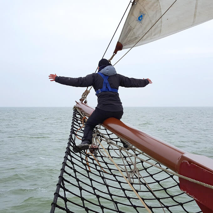 Eine Person sitzt an der Bugspitze eines Segelbootes, unter ihr ein Netz und breitet die Arme aus