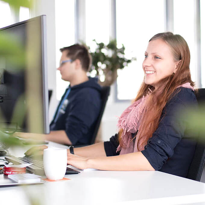 Eine Mitarbeiterin lächelt und blickt auf einen Bildschirm, im Hintergrund ein weiterer Mitarbeiter