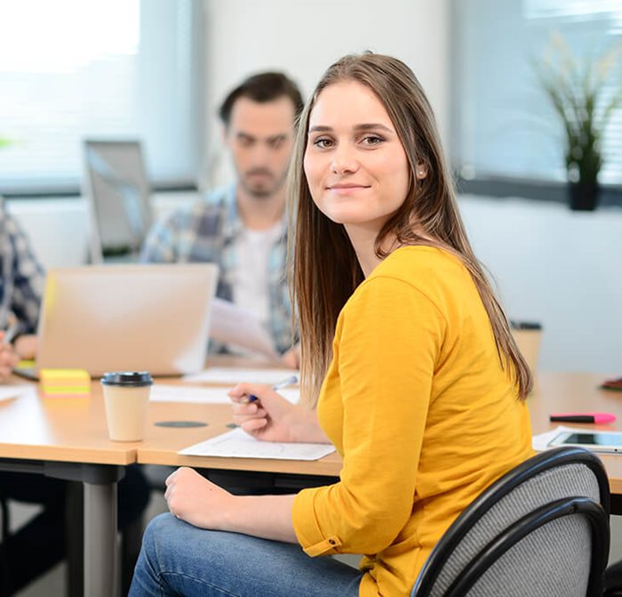 Eine Frau hat ein gelbes Oberteil an und lächelt in die Kamera. Im Hintergrund sitzt ein Mitarbeiter am Schreibtisch.