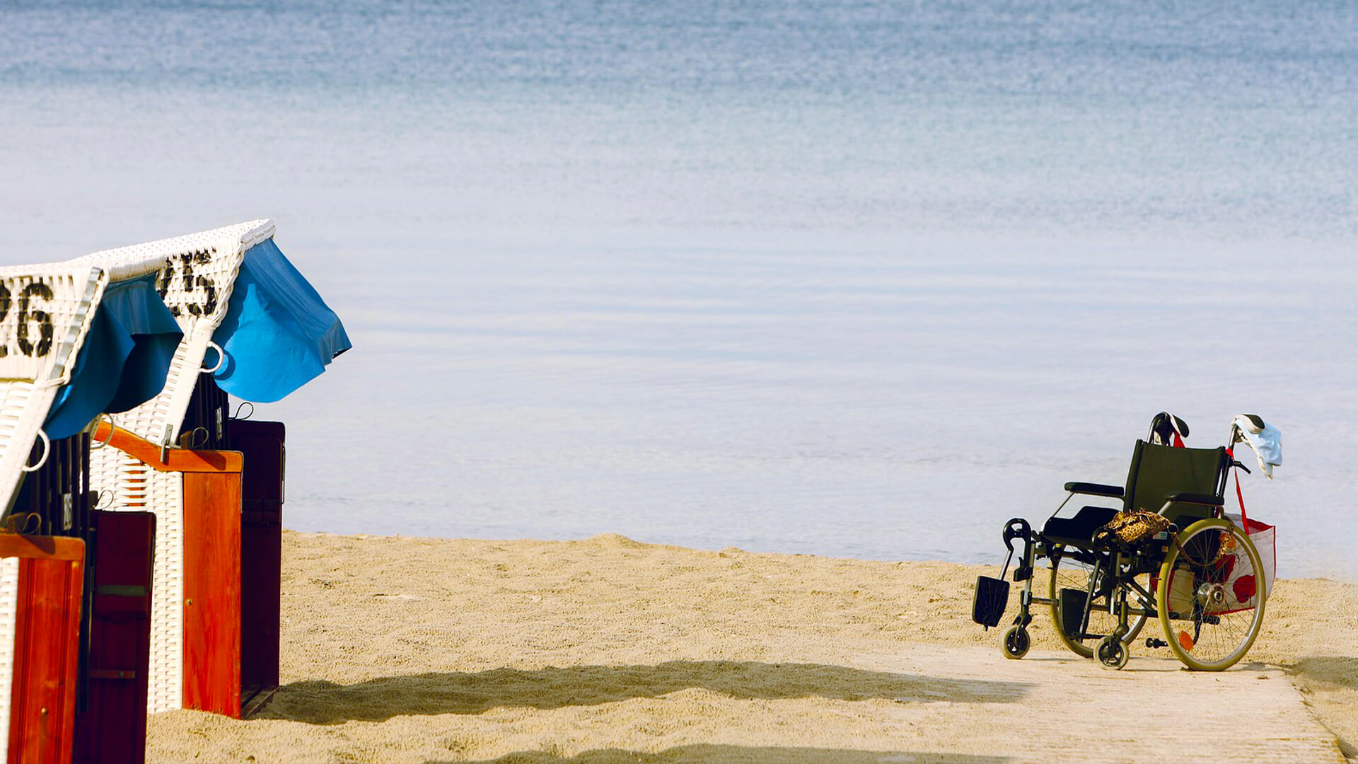 Blick auf einen Strand mit Strandkorb und einem Rollstuhl im Sand.