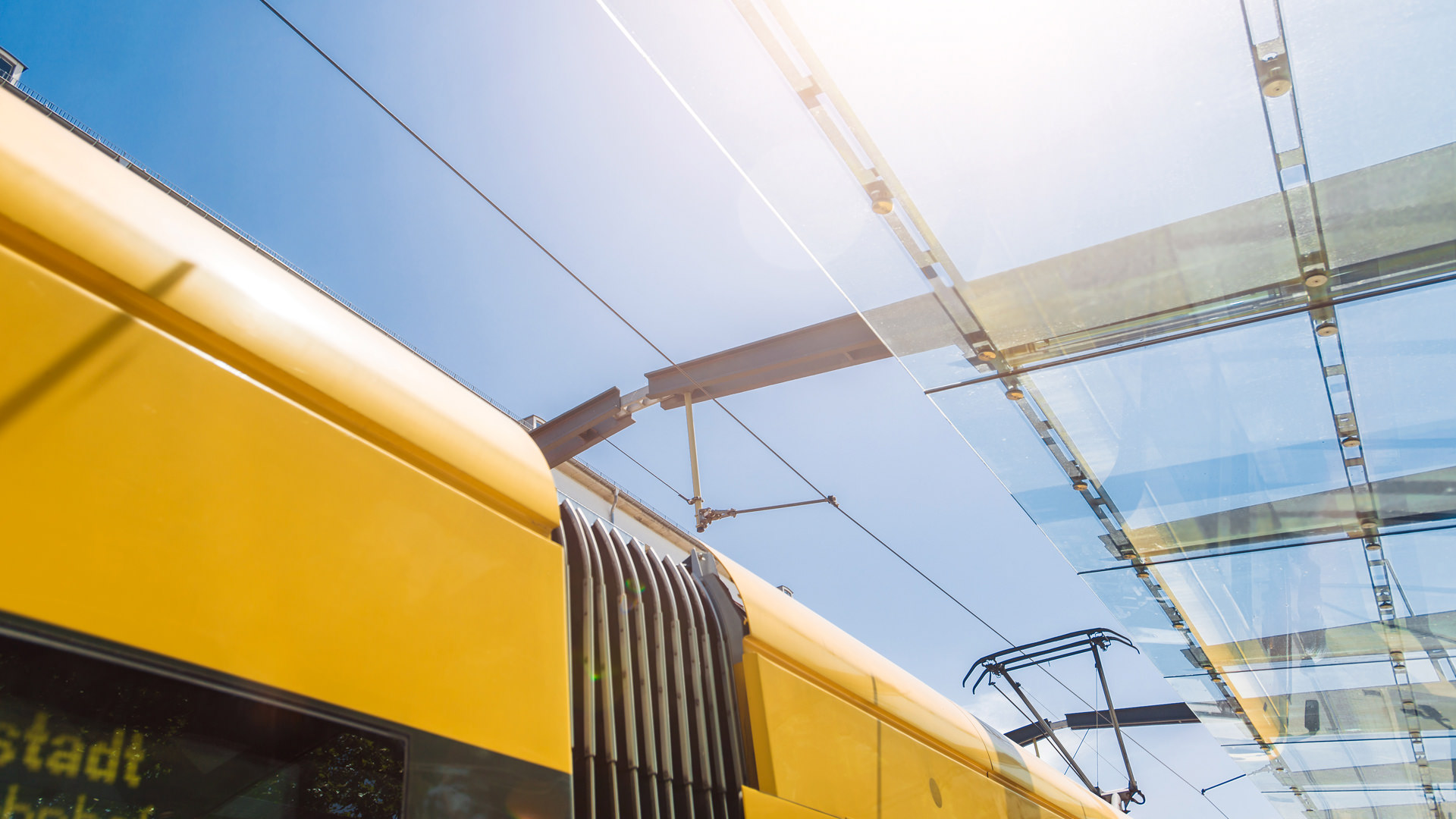 Blick durch die gläserne Bedachung einer Haltestelle Richtung Sonne, links ist im Anschnitt eine gelbe Straßenbahn zu sehen