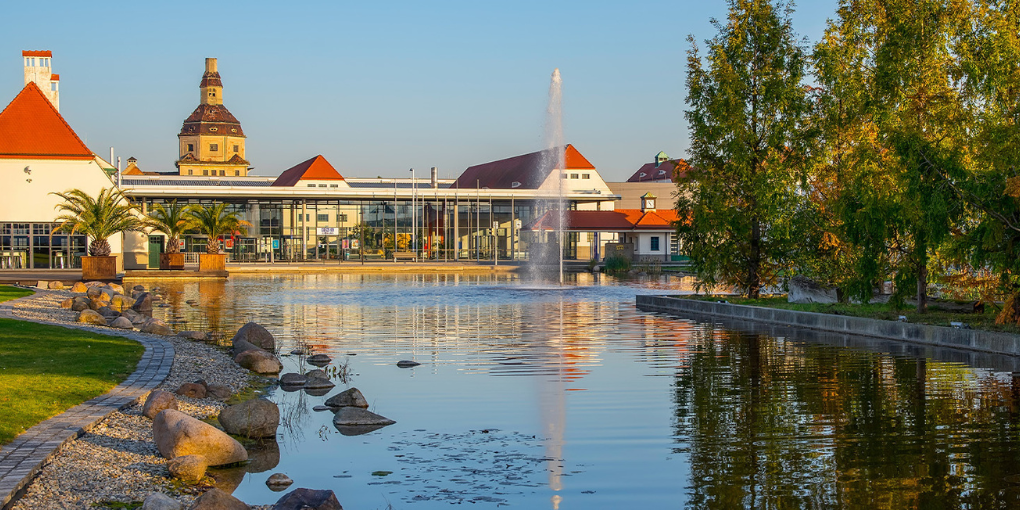 Malerischer Blick auf einen Teich mit einem Springbrunnen, umgeben von Bäumen und Gebäuden mit roten Dächern unter einem klaren blauen Himmel.