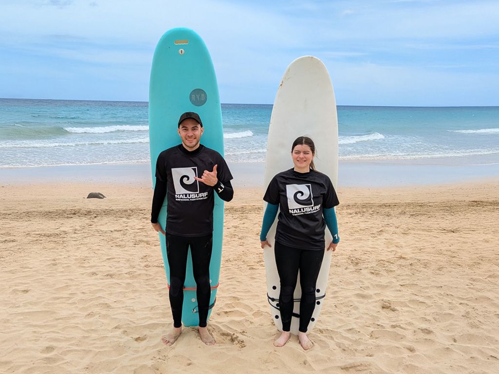 Zwei Personen in Neoprenanzügen stehen an einem Sandstrand, jeweils mit einem Surfbrett in der Hand, im Hintergrund das Meer.