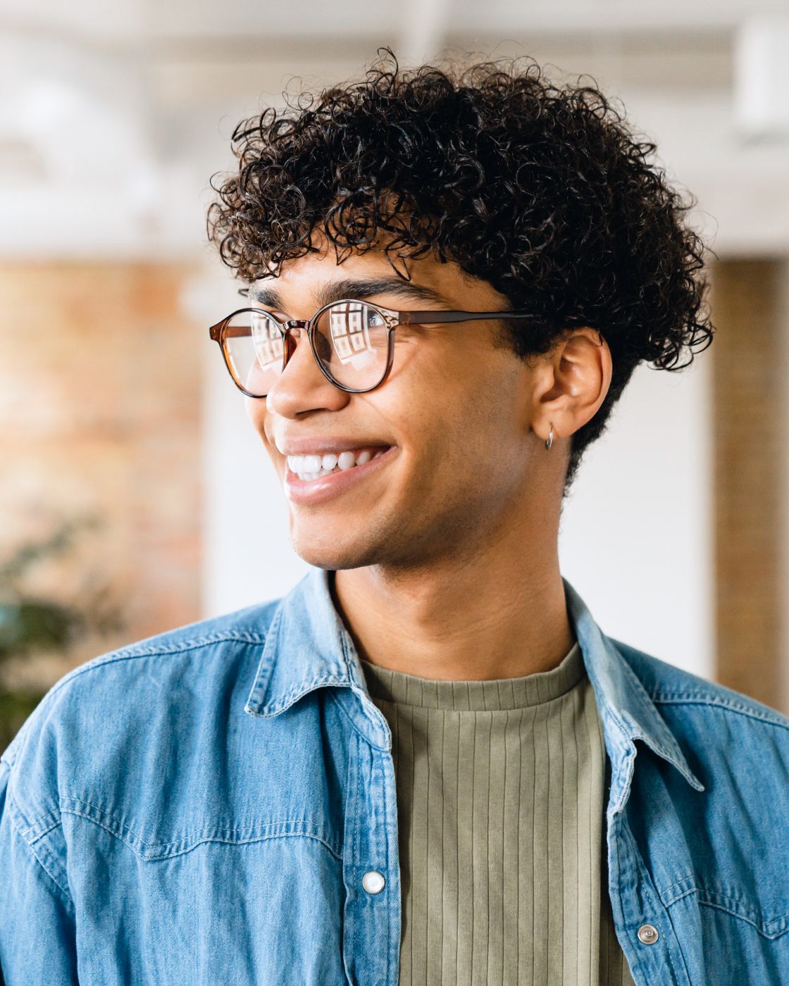 Lächelnde Person mit lockigem Haar und Brille, trägt ein Jeanshemd über einem grünen Oberteil und blickt zur Seite in einem hellen Raum.