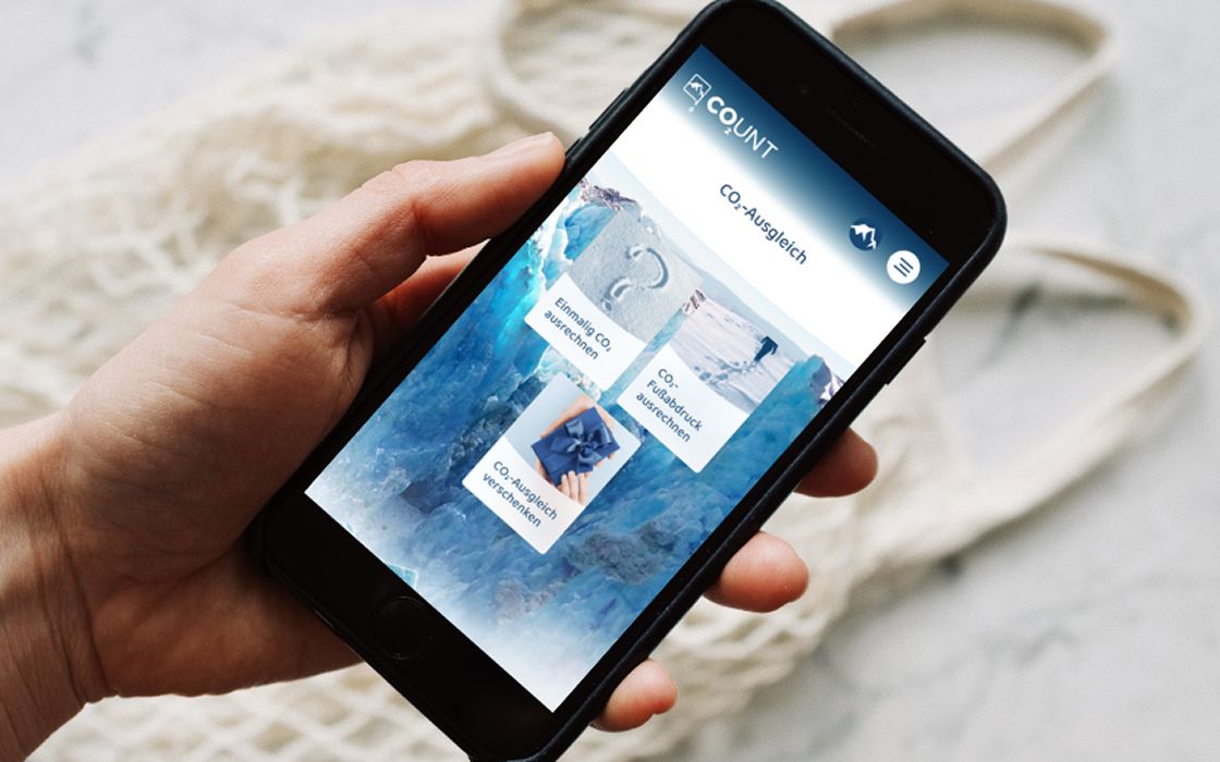Ein Smartphone in der Hand zeigt die App „CO₂unt“ mit verschiedenen Optionen zum CO₂-Ausgleich: CO₂-Fußabdruck berechnen, einmaligen CO₂-Ausgleich durchführen oder CO₂-Ausgleich verschenken. Der Hintergrund zeigt Gletscher-Eis in Blautönen.