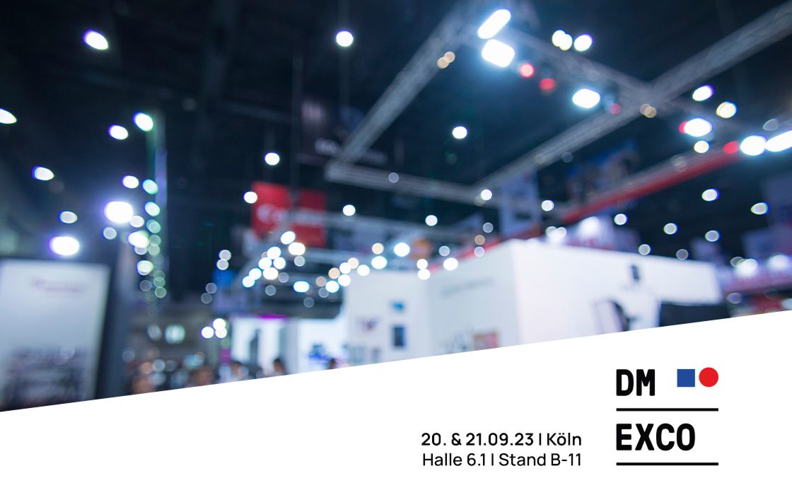 Verschwommene Aufnahme einer Messehalle mit vielen Lichtern und Messeständen. Im Vordergrund steht der Hinweis auf die DMEXCO am 20. und 21.09.2023 in Köln, Halle 6.1, Stand B-11, zusammen mit dem Logo der Messe.