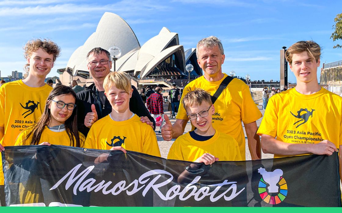 Eine Gruppe von Jugendlichen und zwei Erwachsenen in gelben T-Shirts mit dem Aufdruck „2023 Asia Pacific Open Championship“ posiert mit einem Banner „ManosRobots“ vor dem ikonischen Sydney Opera House.