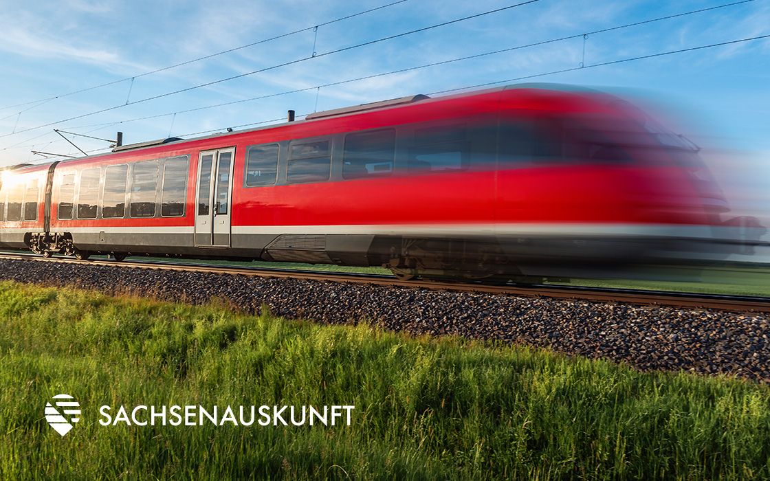 Ein roter Zug fährt mit hoher Geschwindigkeit über Gleise durch ein grasbewachsenes Feld unter einem klaren blauen Himmel. In der Ecke befindet sich das Logo „Sachsenauskunft“.