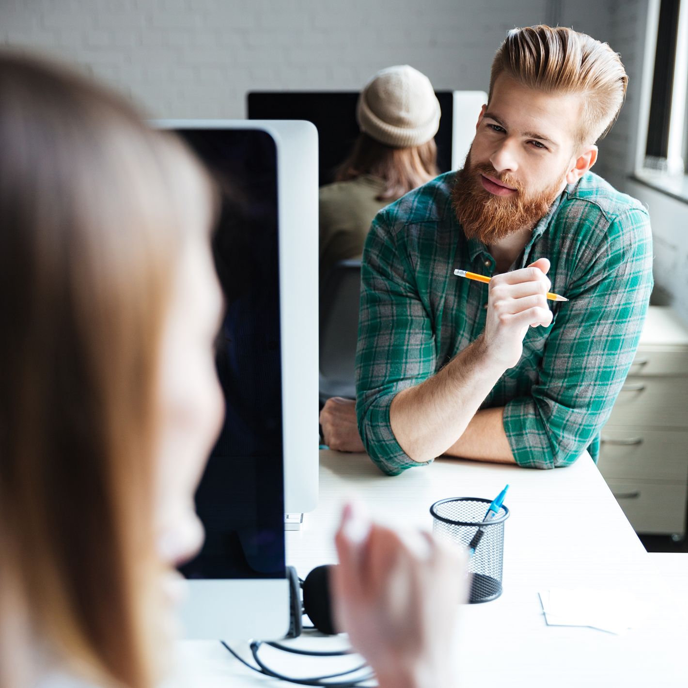 Junger Mann mit rotem Bart und grün karierten Hemd hält einen Stift in der Hand und hört aufmerksam einer Kollegin zu, die ihm gegenüber sitzt.