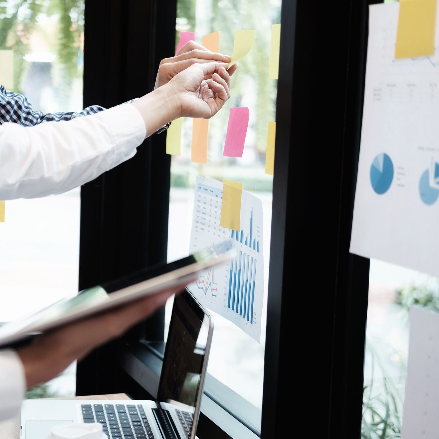 Menschen arbeiten gemeinsam an einem Fenster mit Haftnotizen und Diagrammen und nutzen dabei einen Laptop. Helles, natürliches Licht erhellt den Arbeitsbereich.
