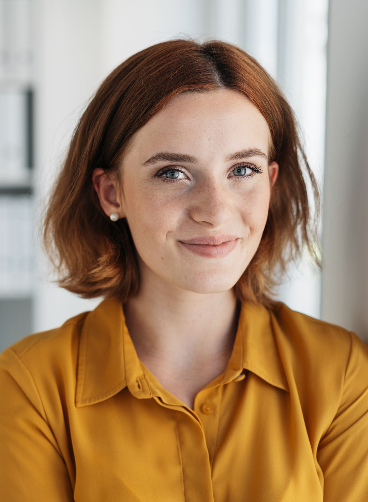 Lächelnde Frau mit kurzem kastanienbraunem Haar, trägt eine senfgelbe Bluse und steht in einem gut beleuchteten Raum mit neutralem Hintergrund.
