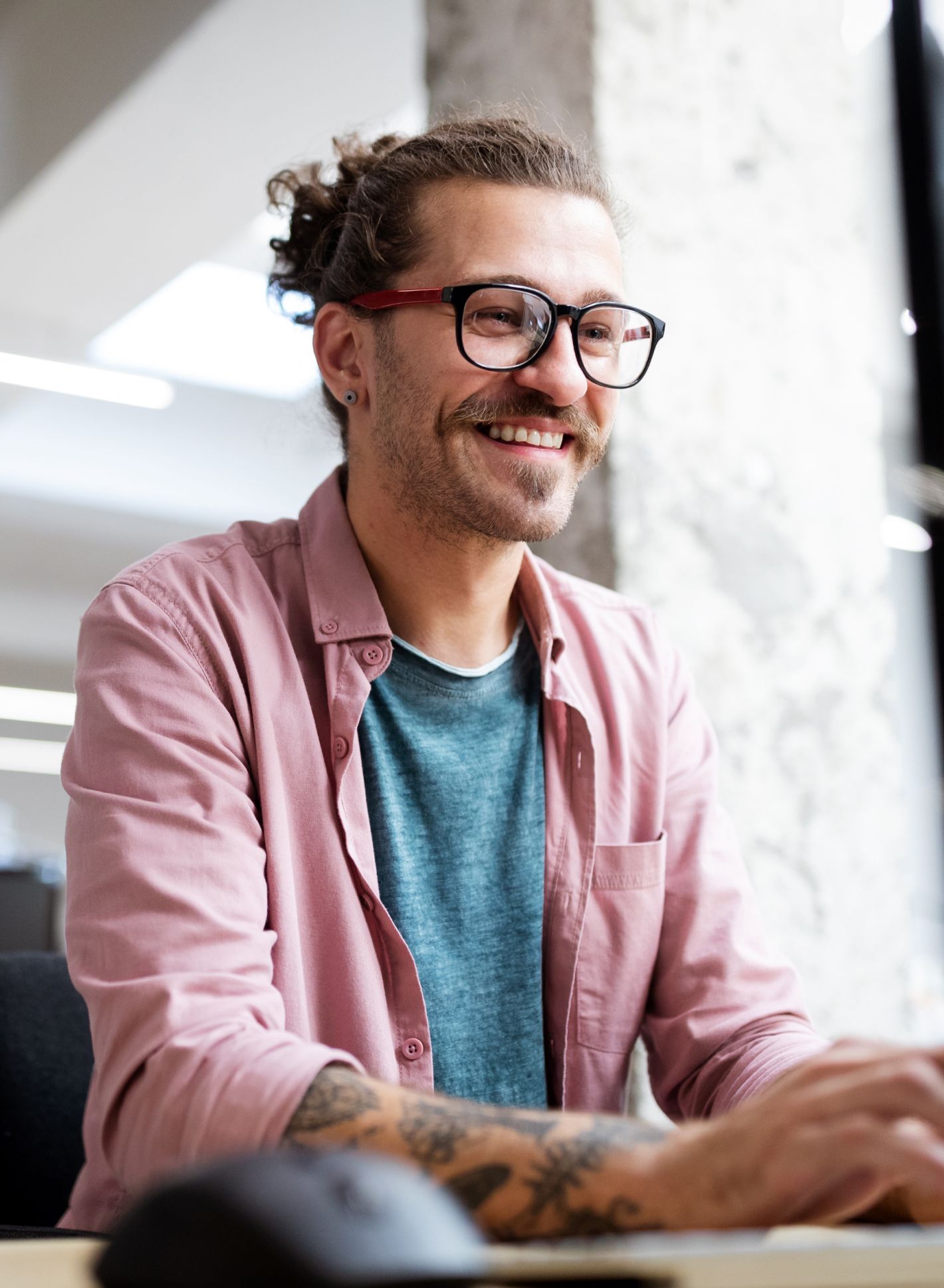 Lächelnde Person mit Brille und Schnurrbart, trägt ein pinkes Hemd über einem türkisfarbenen Oberteil, sitzt und arbeitet an einem Computer in einem hellen Raum.