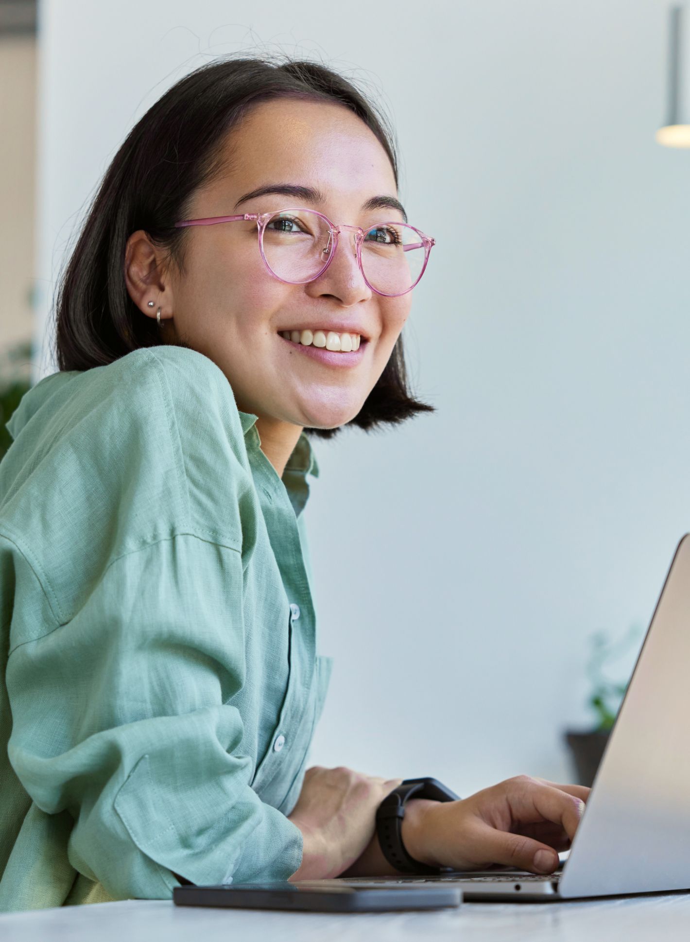 Lächelnde Person mit Brille und grünem Shirt arbeitet an einem Laptop und sitzt an einem Schreibtisch, auf dem ein Smartphone in der Nähe liegt.