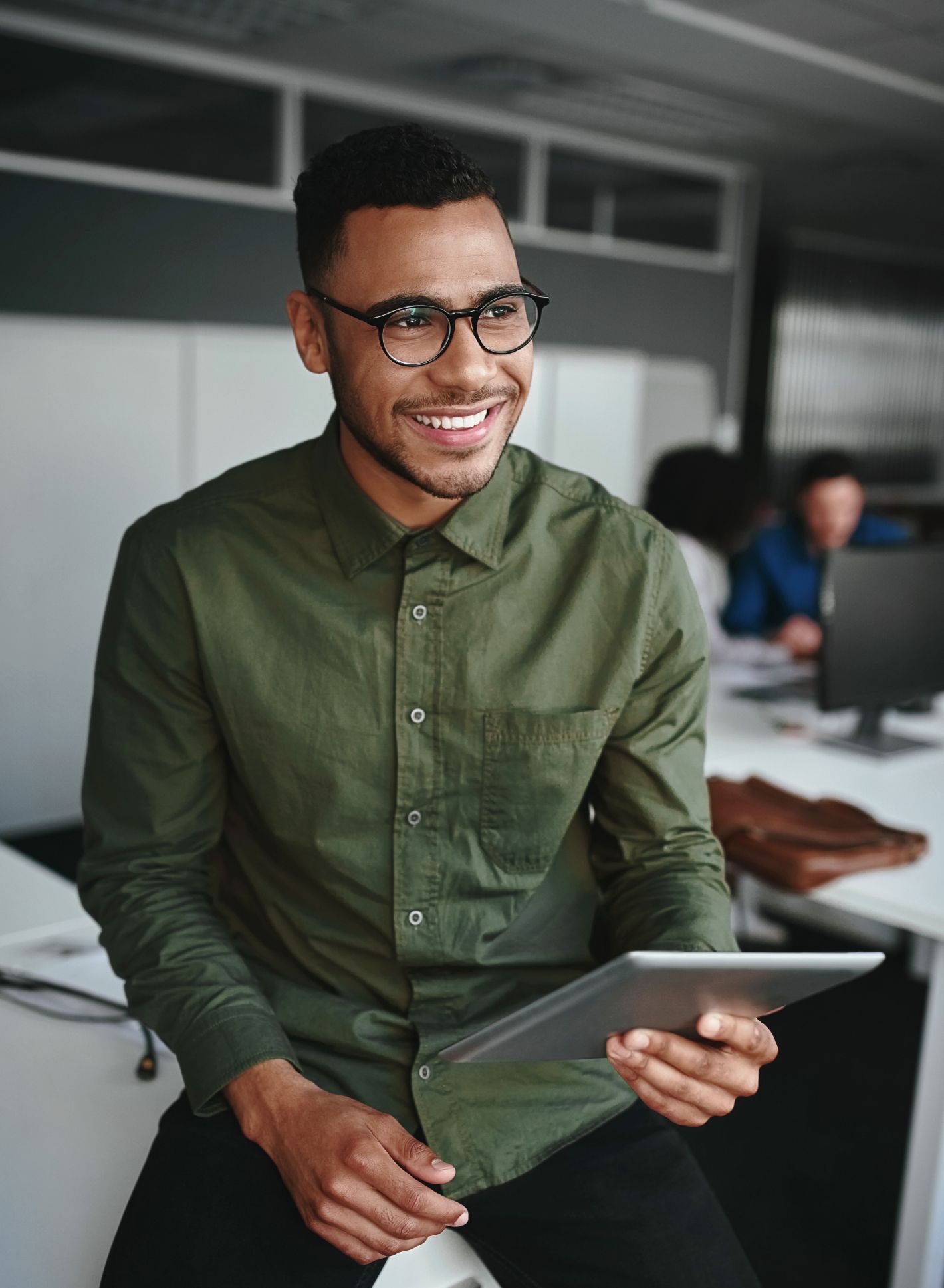 Lächelnder Mann mit Brille und grünem Hemd hält ein Tablet und sitzt in einem modernen Büro, während im Hintergrund Kollegen arbeiten.