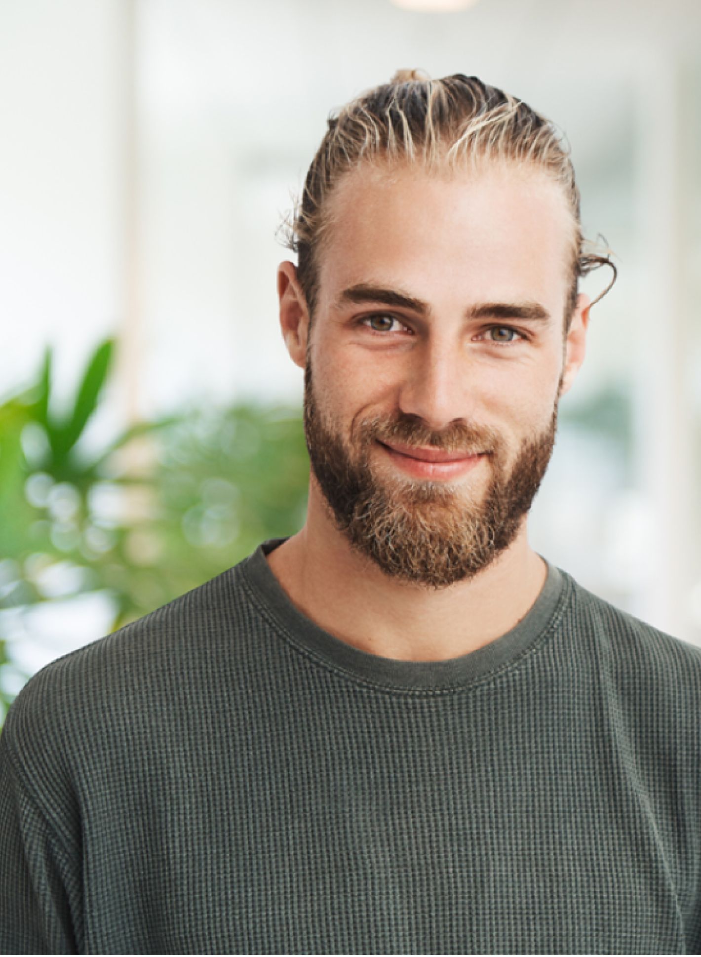 Lächelnder Mann mit Bart und zurückgebundenem Haar, trägt einen grünen Pullover und steht in einem Innenraum mit verschwommenem Grün im Hintergrund.