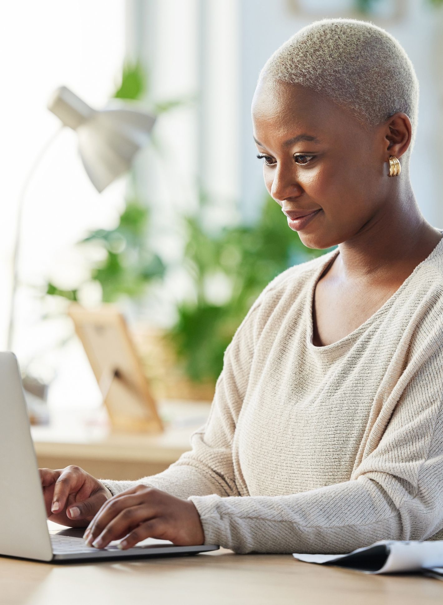 Eine Person mit kurzen Haaren tippt an einem Laptop an einem Schreibtisch, umgeben von Pflanzen und einer Lampe, in einem hellen Raum.