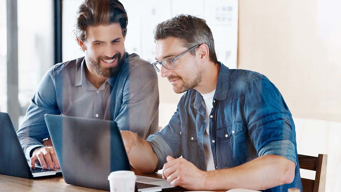 Zwei Männer arbeiten gemeinsam an einem Tisch mit Laptops, einer zeigt auf den Bildschirm. Sie wirken engagiert und konzentriert in einem hellen Büro.