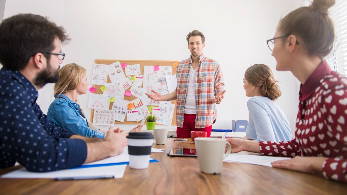 Ein Mann in kariertem Hemd hält eine Präsentation vor einem Team (4 weitere Personen) in einem kreativen Büro. Die Kollegen sitzen an einem Tisch und hören ihm aufmerksam zu, während eine Pinnwand mit Diagrammen und Notizen im Hintergrund zu sehen ist.