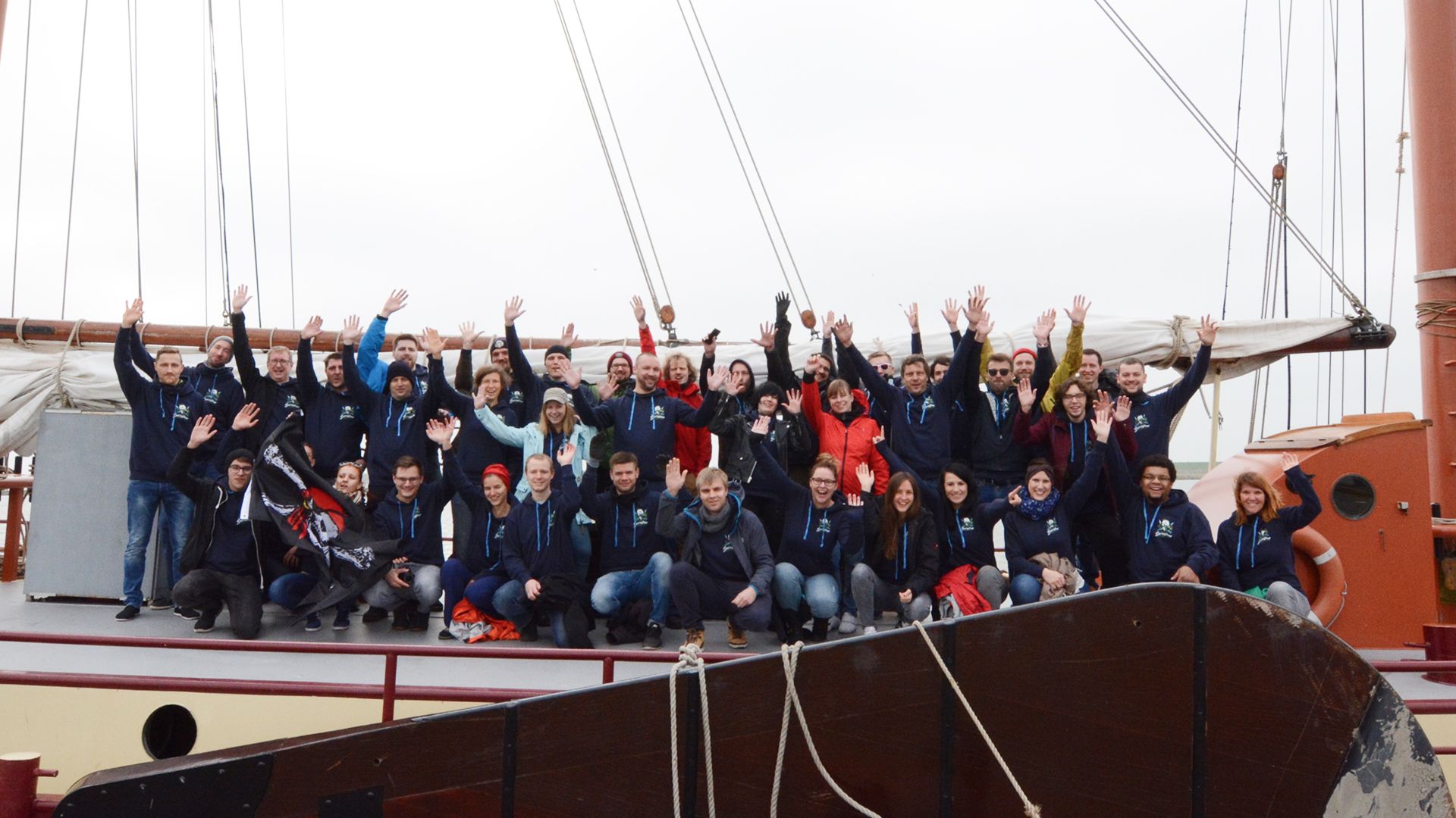 Eine große Gruppe von Personen posiert gemeinsam auf einem Boot, alle lächeln und heben jubelnd die Hände.