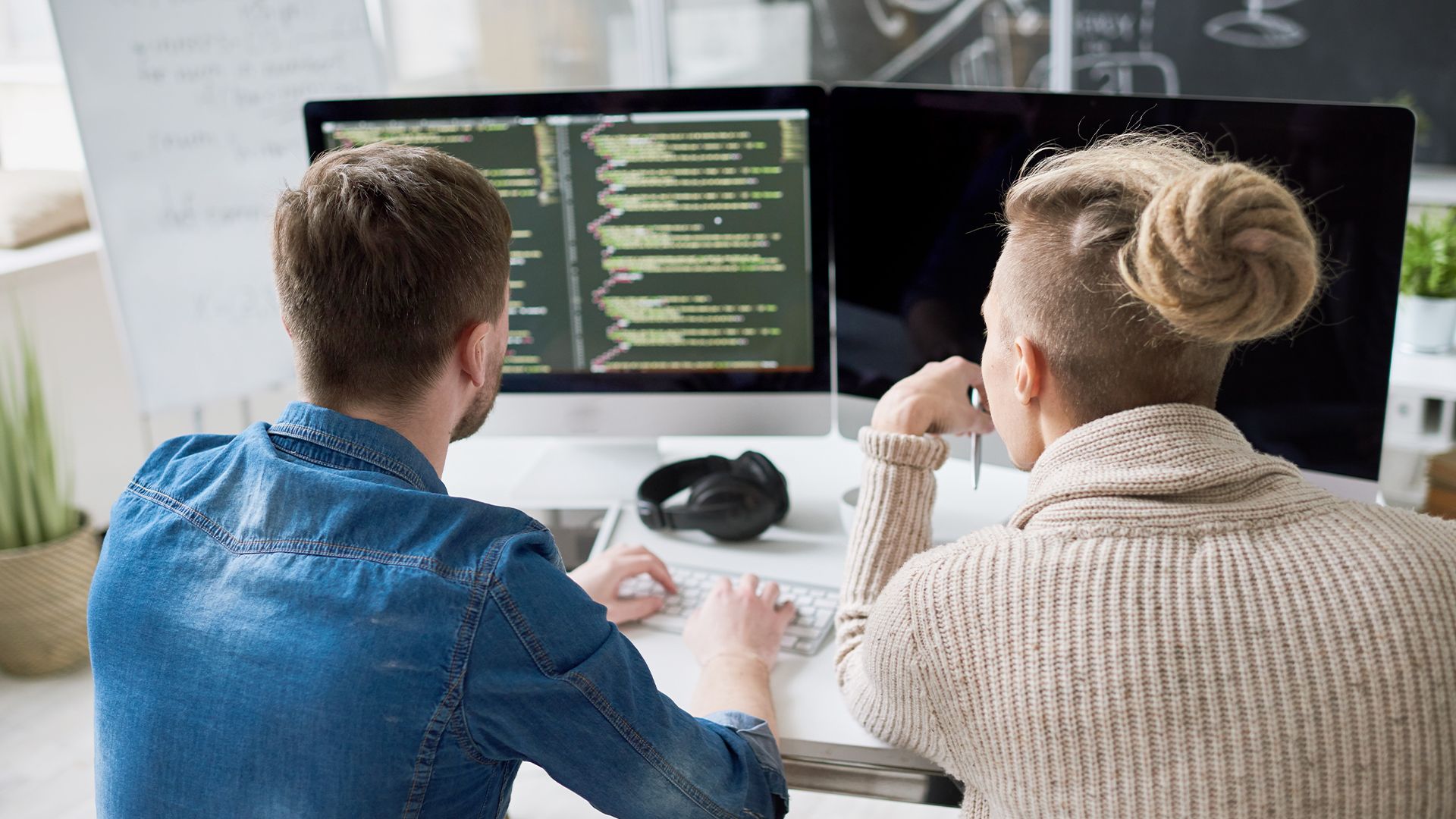 Zwei Personen sitzen an einem Schreibtisch und betrachten Code auf zwei Computermonitoren. Eine hat die Haare zu einem Dutt gebunden, die andere trägt ein Jeanshemd.
