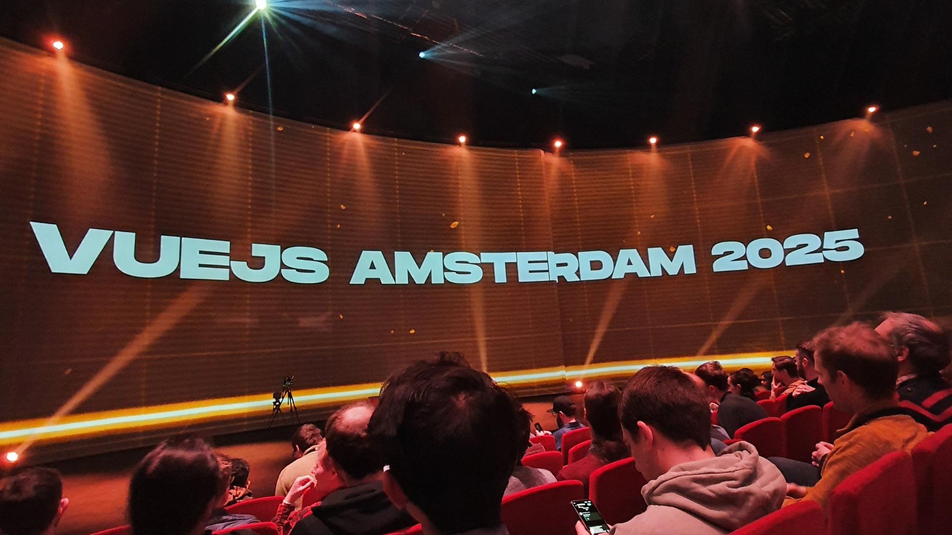 Publikum sitzt in einem Theater, während „VUEJS AMSTERDAM 2025“ auf eine große, gebogene Leinwand projiziert wird.