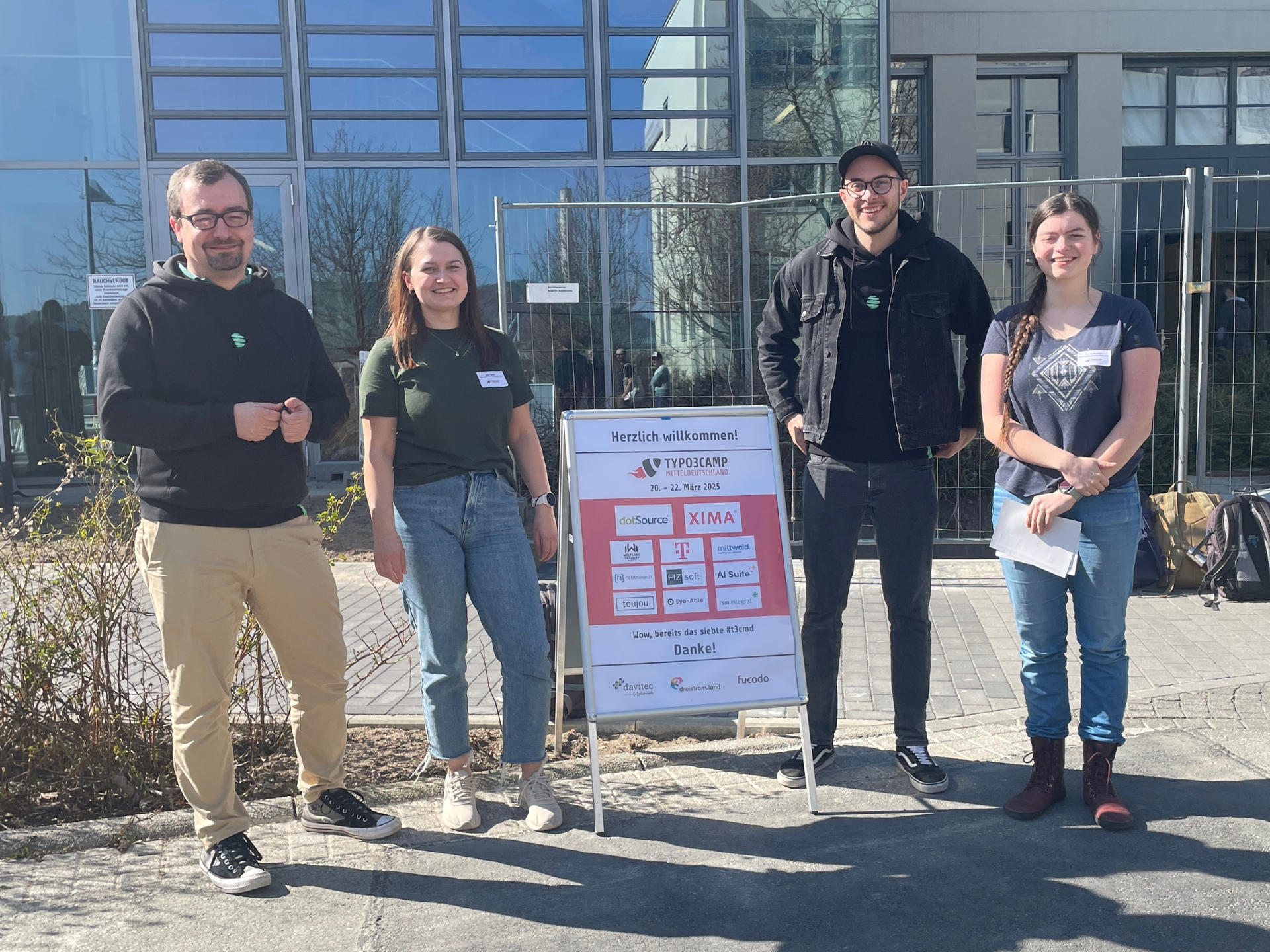 Vier Personen stehen lächelnd im Freien nahe einem Willkommensschild vor einem modernen Gebäude mit großen Fenstern.