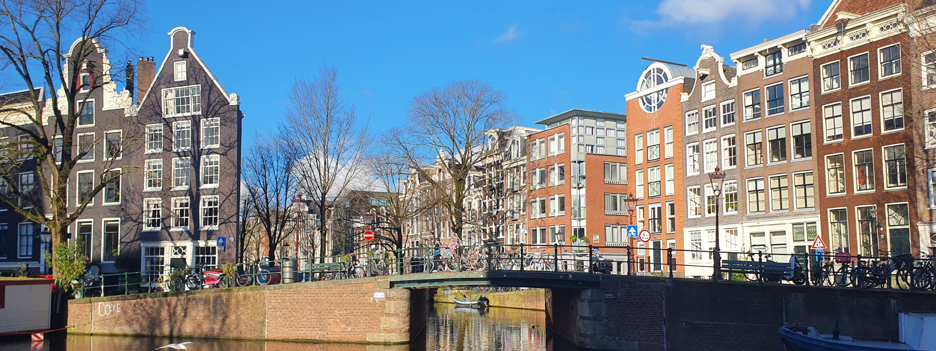 Ein sonniger Blick auf Amsterdams Kanal, gesäumt von traditionellen niederländischen Gebäuden, einer Brücke und kahlen Bäumen unter einem klaren blauen Himmel.