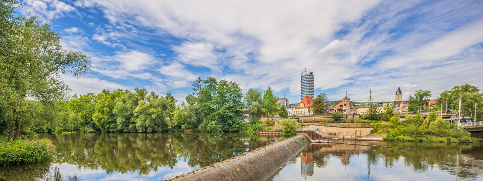 Malerischer Blick auf einen Fluss mit üppigem Grün, einer kleinen Stadt und einem hohen Gebäude unter einem teilweise bewölkten blauen Himmel.