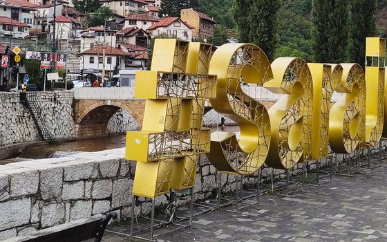 Eine große goldene 3D-Installation mit der Aufschrift „#Sarajevo“ steht auf einer Promenade entlang eines Flusses. Im Hintergrund sind traditionelle Gebäude mit roten Dächern und eine Steinbrücke über den Fluss zu sehen. Umgeben von Hügeln und Bäumen vermittelt die Szene ein Gefühl von historischer und moderner Kultur.