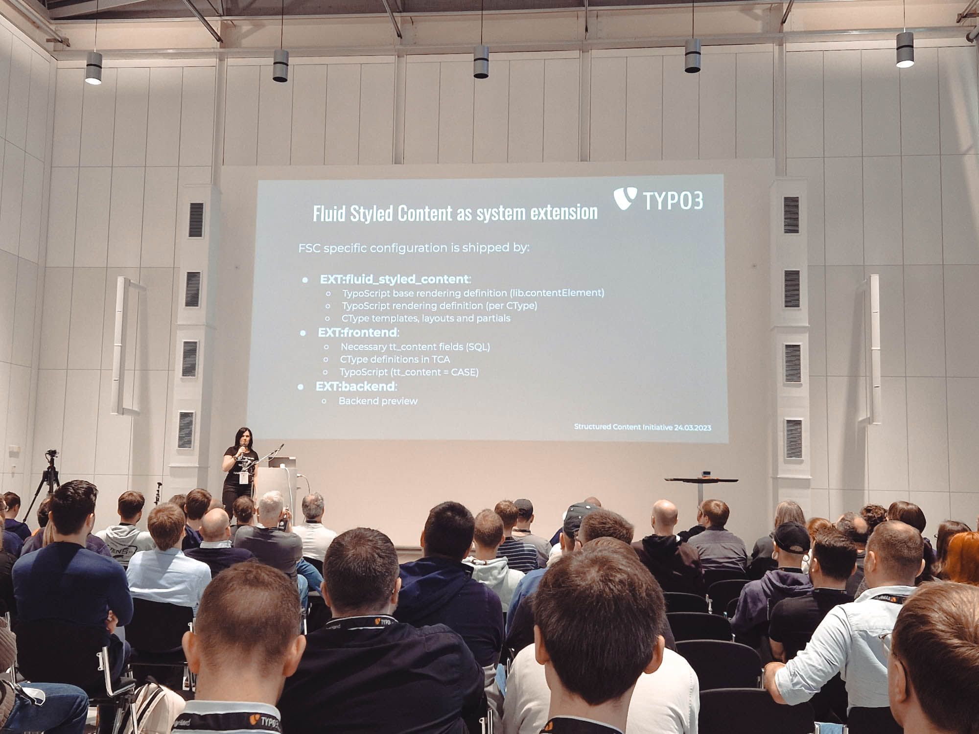 Eine Vortragssituation in einem modernen Konferenzsaal. Im Vordergrund sitzt ein Publikum, während auf einer großen Leinwand eine Präsentation mit dem Titel „Fluid Styled Content as system extension“ gezeigt wird. Eine Person steht links neben der Leinwand am Rednerpult und spricht. Die Folie listet Konfigurationspunkte zu TYPO3-Extensions auf.