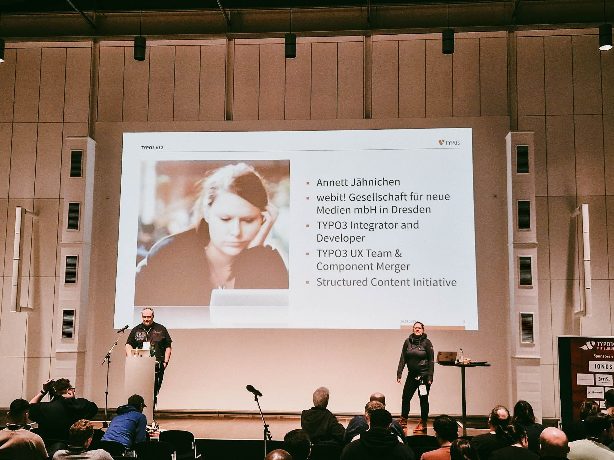 Ein Vortrag in einem Konferenzsaal. Zwei Personen stehen auf der Bühne vor einer großen Leinwand. Auf der Leinwand wird ein Bild von Annett Jähnichen sowie Informationen über ihre Rolle bei TYPO3 und der Structured Content Initiative angezeigt. Vor der Bühne sitzt ein Publikum, das der Präsentation aufmerksam folgt.