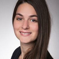 Lächelnde Frau mit langen braunen Haaren, die einen dunklen Blazer trägt und vor einem neutralen Hintergrund posiert.