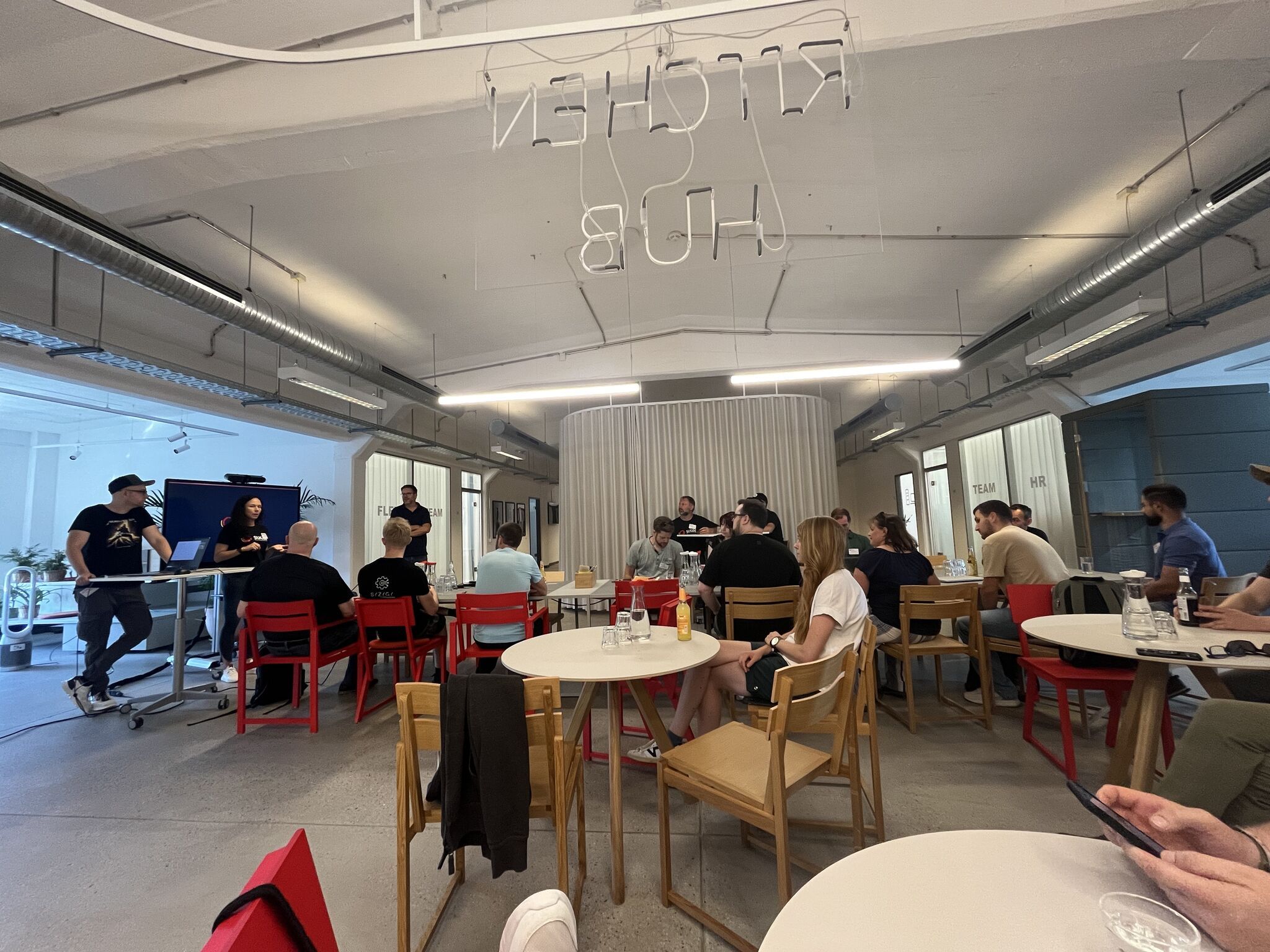 Eine Gruppe von Personen sitzt in einem modernen Besprechungsraum mit runden Tischen und verfolgt eine Präsentation.