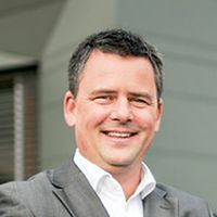 Lächelnder Mann im grauen Anzug mit kurzen dunklen Haaren, steht im Freien vor einem modernen Gebäude.