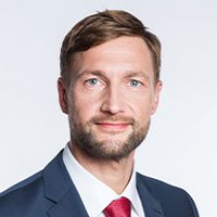 Ein Mann mit kurzen braunen Haaren und Bart trägt einen blauen Anzug, ein weißes Hemd und eine rote Krawatte vor einem schlichten weißen Hintergrund.