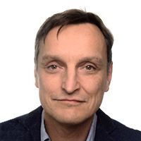 Ein Mann mit kurzen dunklen Haaren und einem leichten Lächeln trägt eine dunkle Jacke über einem hellen Hemd, vor einem schlichten weißen Hintergrund.