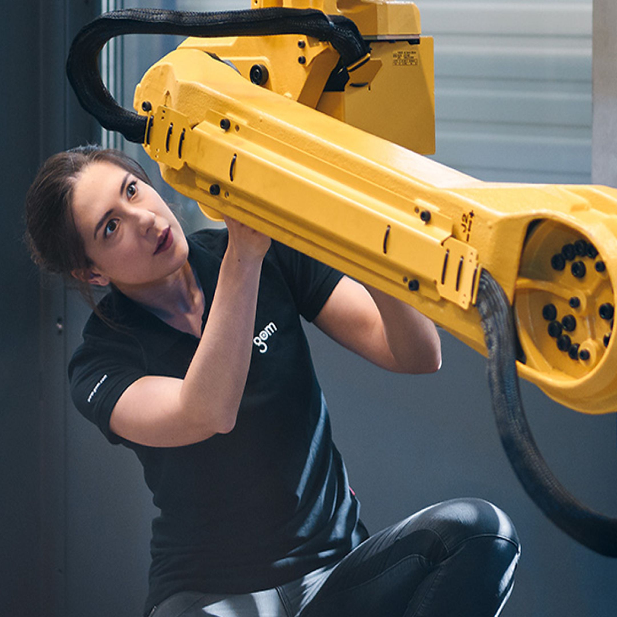 Eine Frau in einem schwarzen Shirt inspiziert einen großen gelben Industrieroboterarm in einer Werkstattumgebung.