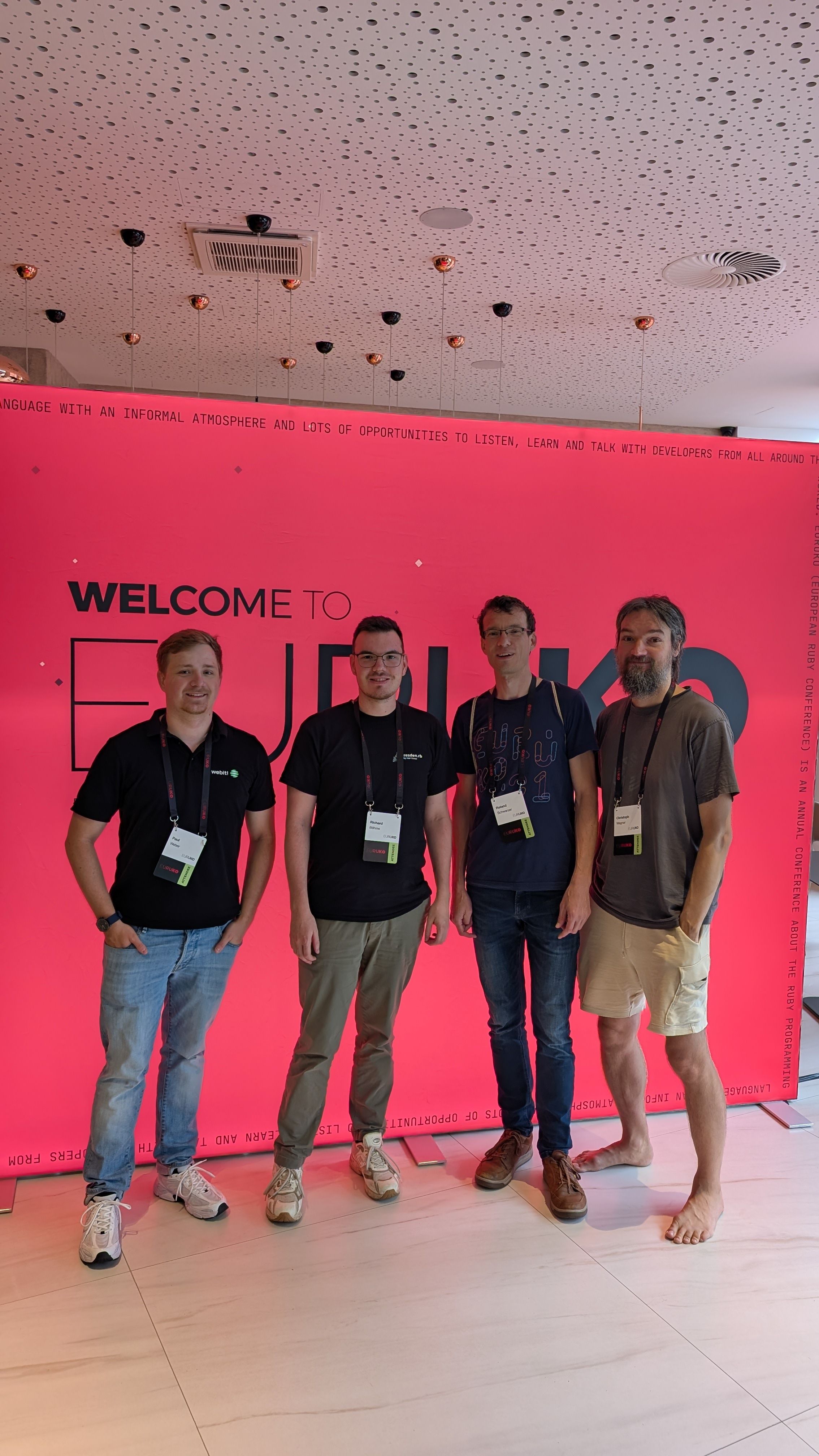 Vier Männer stehen zusammen vor einer leuchtend rosa Wand, auf der „Welcome to EuRuKo“ steht. Sie lächeln und tragen Freizeitkleidung.