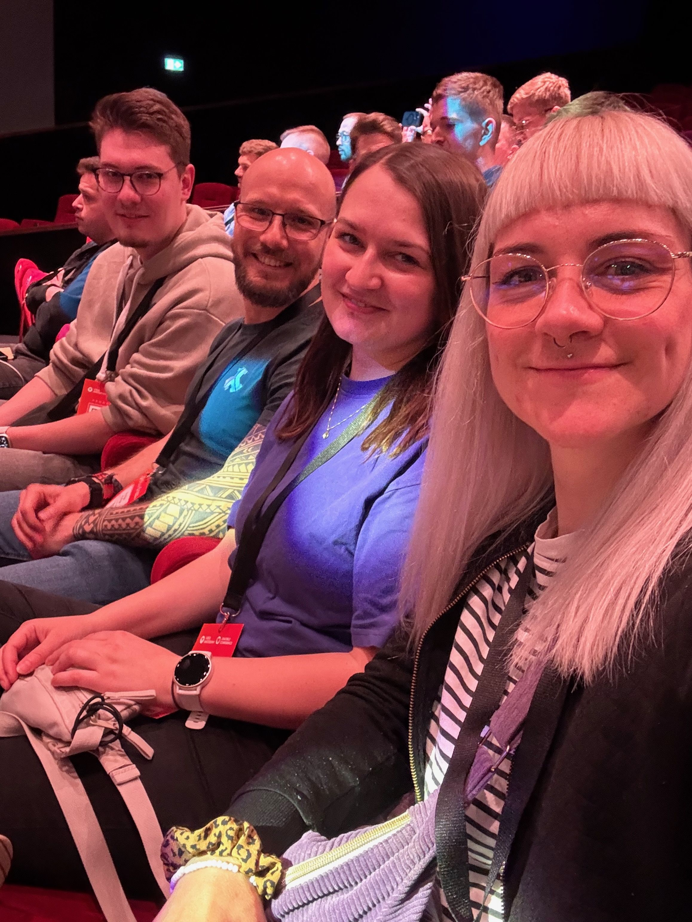 Eine Gruppe von Personen sitzt in einem Theater und lächelt in die Kamera. Sie tragen legere Kleidung und haben Veranstaltungs-Lanyards um den Hals.