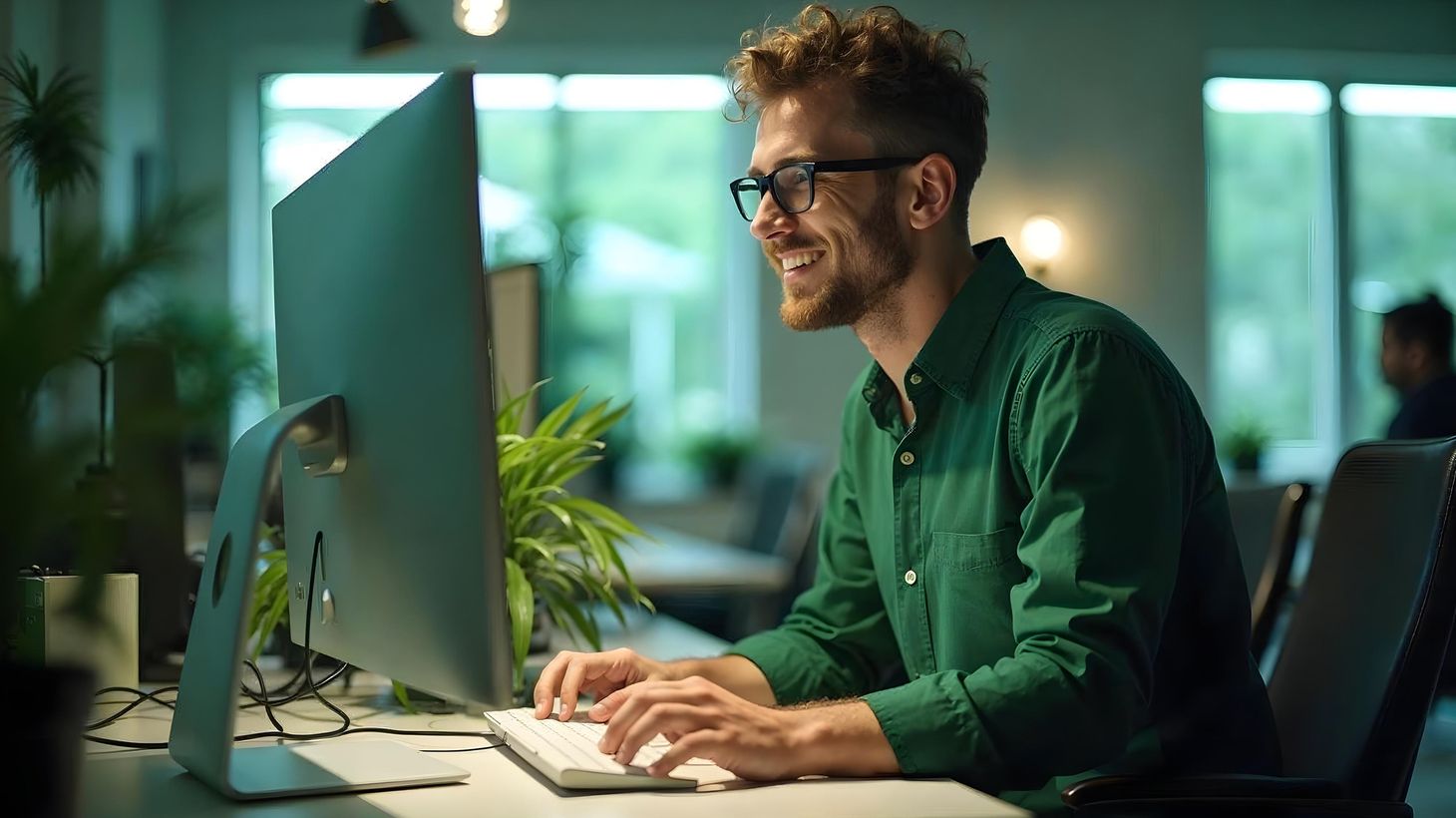 Ein Mann mit Brille und grünem Hemd lächelt, während er an einem Computer in einem modernen Büro mit Pflanzen und großen Fenstern arbeitet.
