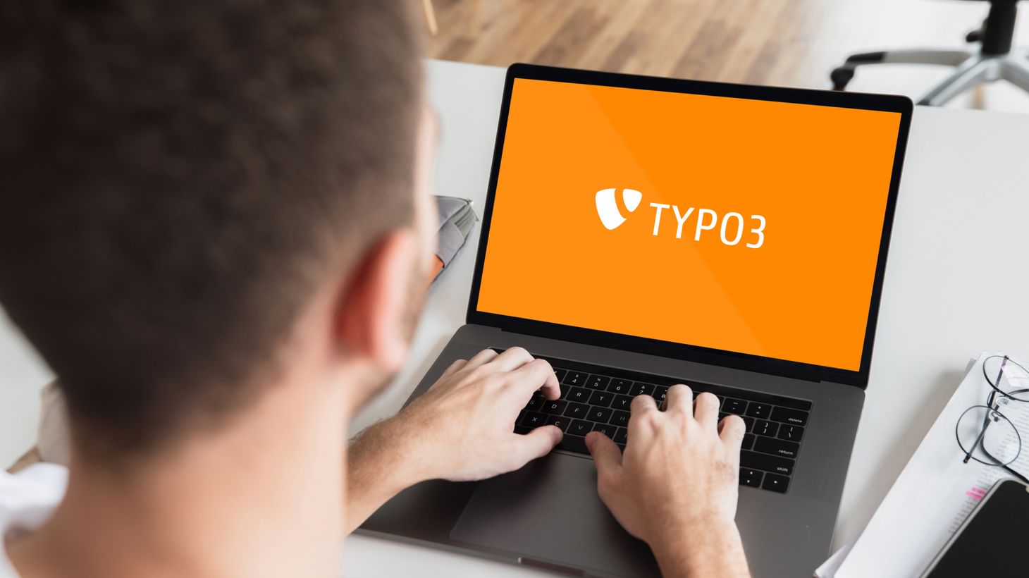 Eine Person nutzt einen Laptop mit dem TYPO3-Logo auf einem orangefarbenen Bildschirm und sitzt an einem Schreibtisch mit unscharfem Hintergrund.