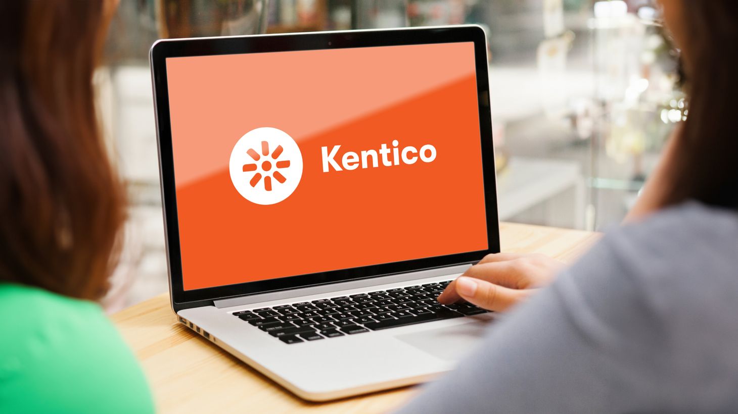 Eine Person nutzt einen Laptop mit dem Kentico-Logo auf einem orangefarbenen Bildschirm und sitzt an einem Holztisch.
