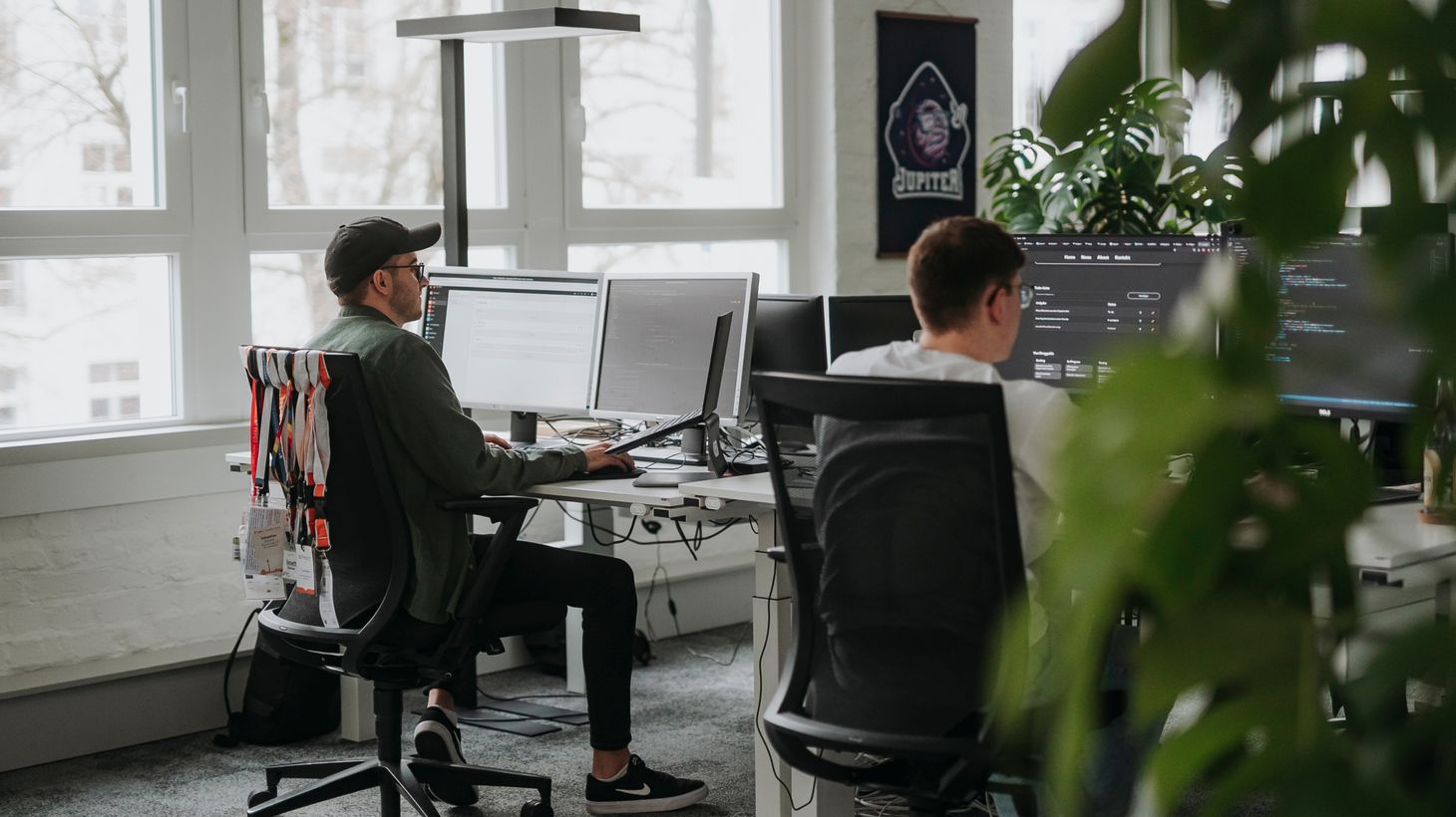 Zwei Personen arbeiten an Schreibtischen mit mehreren Monitoren in einem modernen Büro, umgeben von Grünpflanzen und großen Fenstern.