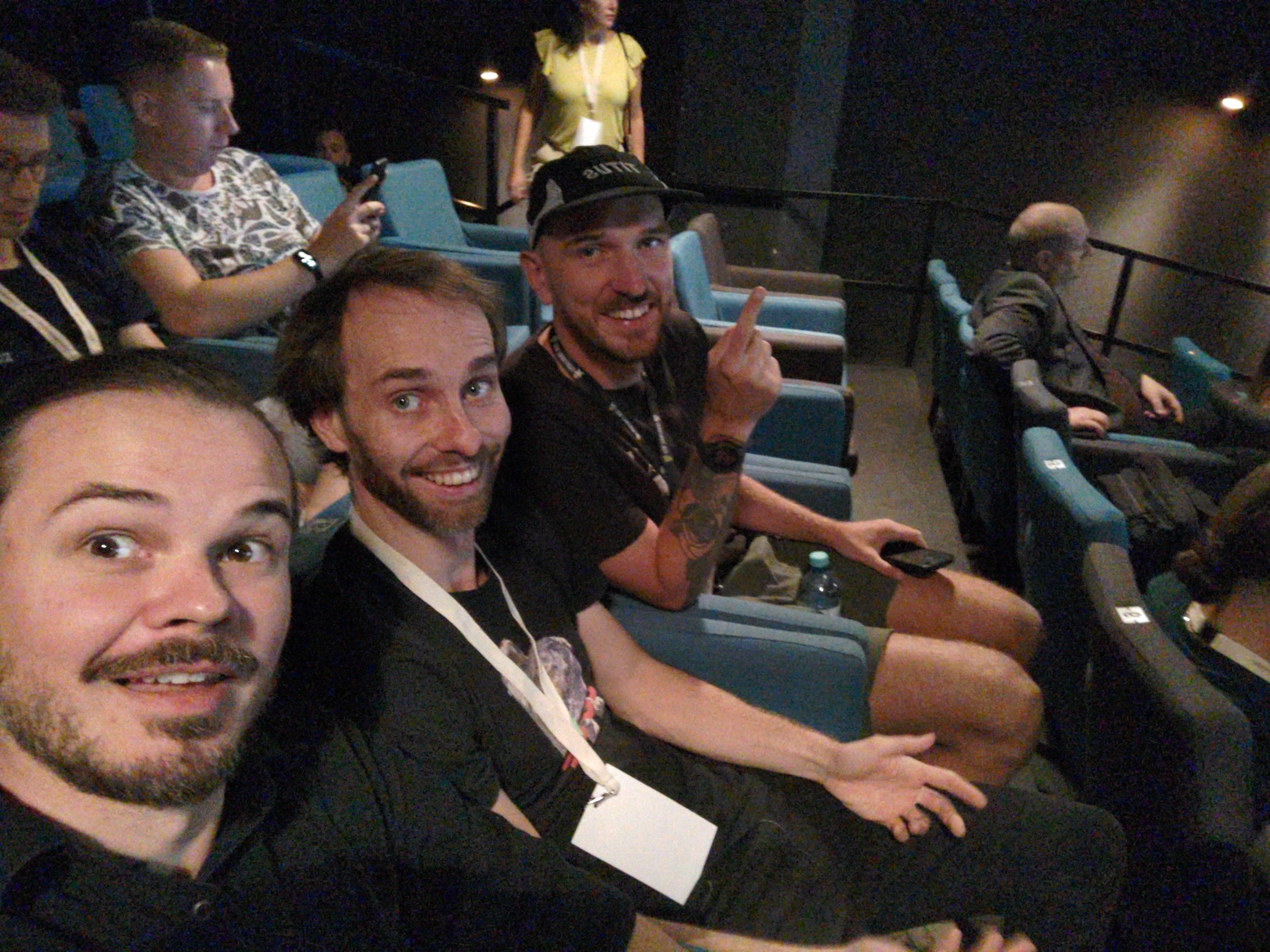 Drei Männer sitzen in einem Kinosaal mit blauen Sesseln. Sie lächeln und posieren für ein Selfie. Einer der Männer trägt eine schwarze Kappe, zeigt mit dem Finger nach oben und hat ein Tattoo am Arm. Im Hintergrund sind weitere Teilnehmer zu sehen, einige schauen auf ihre Smartphones.