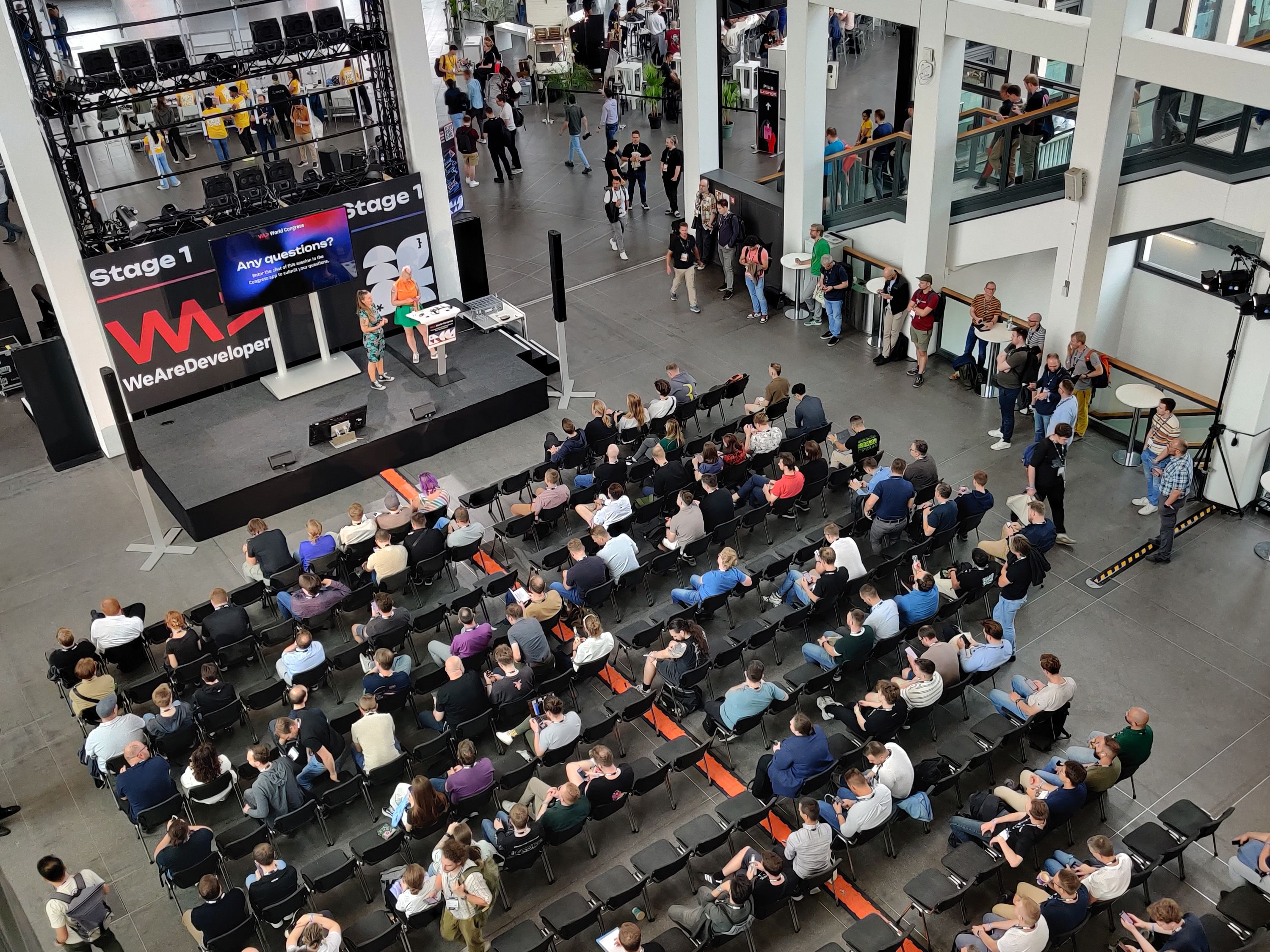 Draufsicht auf Konferenz mit sitzenden Teilnehmern, einem Sprecher auf der Bühne und einer großen Leinwand mit der Aufschrift „WeAreDevelopers“.