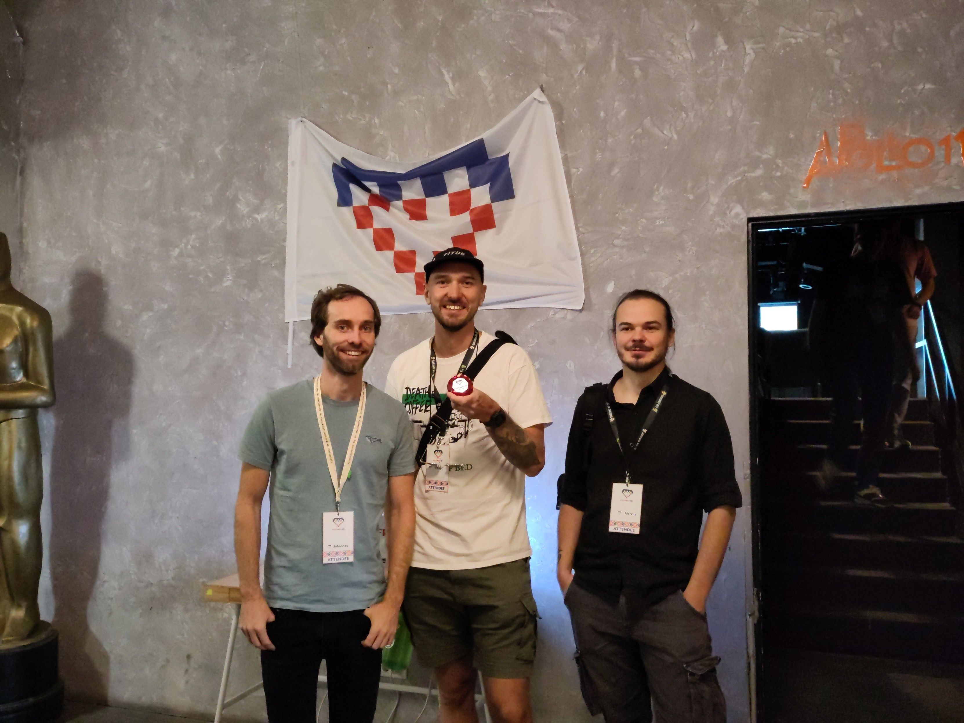 Drei Männer stehen nebeneinander vor einer Betonwand mit einer Flagge, die ein pixeliertes Herzmuster zeigt. Sie tragen Konferenz-Namensschilder und lächeln in die Kamera. Einer von ihnen hält eine kleine Medaille oder einen Button in der Hand. Links neben ihnen steht eine goldene Statue, und rechts führt eine dunkle Treppe nach oben, wo Personen hinaufgehen.