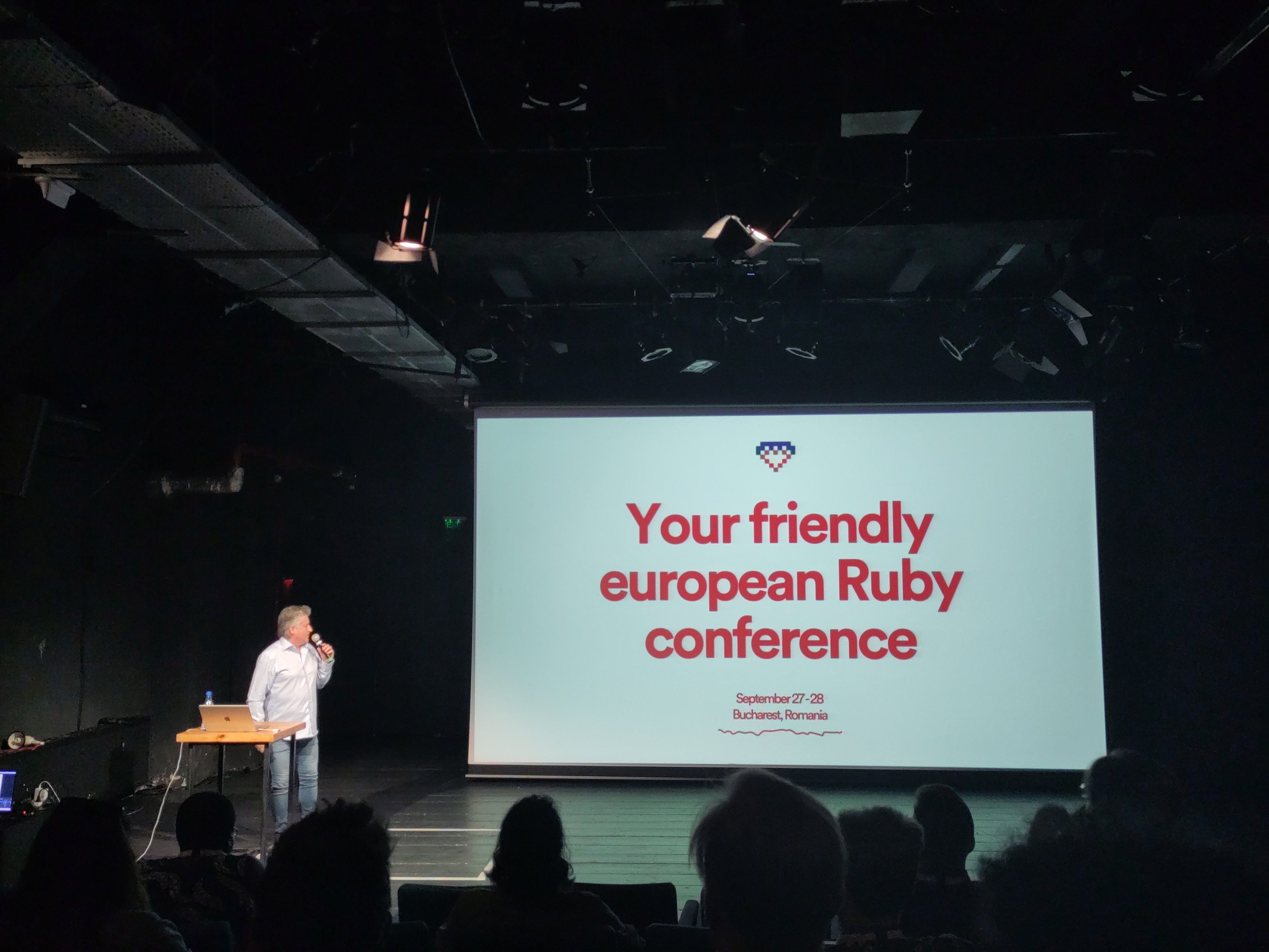 Eine Person auf einer Bühne hält ein Mikrofon und steht neben einem kleinen Tisch mit einem Laptop. Im Hintergrund ist eine große Präsentationsfolie zu sehen, die den Text „Your friendly European Ruby conference“ zeigt, zusammen mit dem Datum „September 27-28, Bucharest, Romania“. Die Umgebung ist ein dunkler Veranstaltungsraum mit Publikumsreihen.