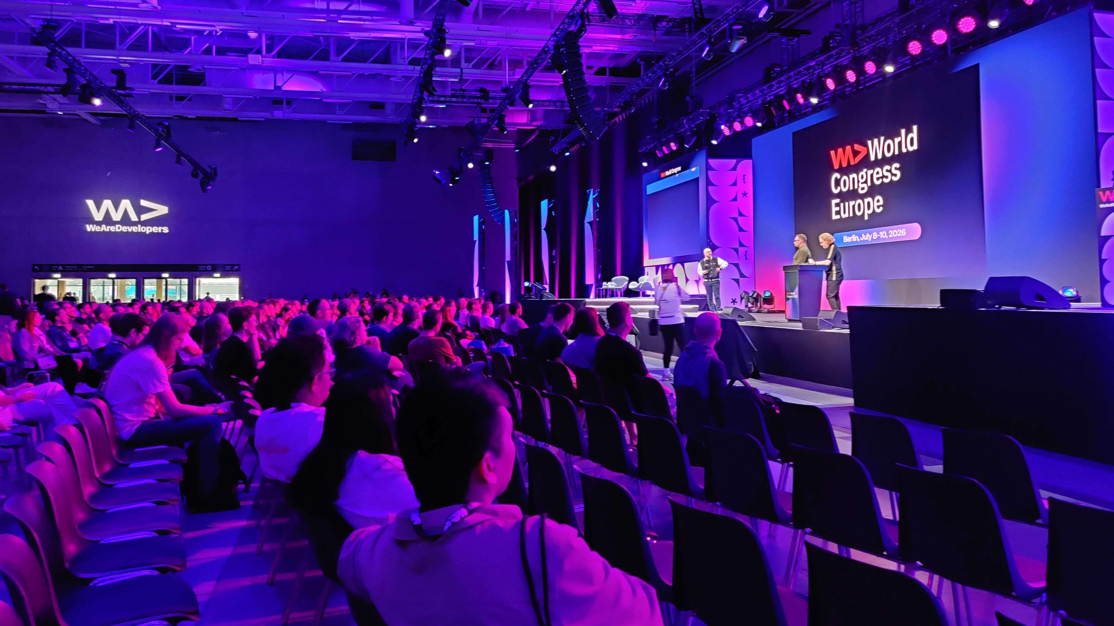Publikum sitzt bei der Konferenz vor einer violett beleuchteten Bühne mit der Aufschrift „World Congress Europe“. Redner stehen an Rednerpulten.